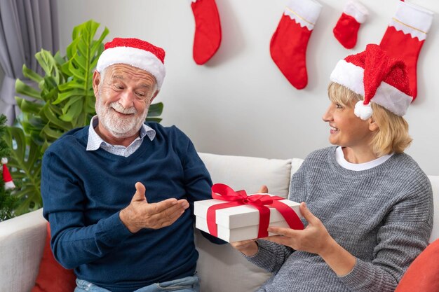 Senior couple husband and wife exchange giving gift present sitting on sofa in room with Christmas tree and decoration