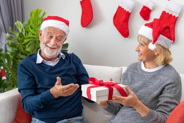 Senior couple husband and wife exchange giving gift present sitting on sofa in room with Christmas tree and decoration