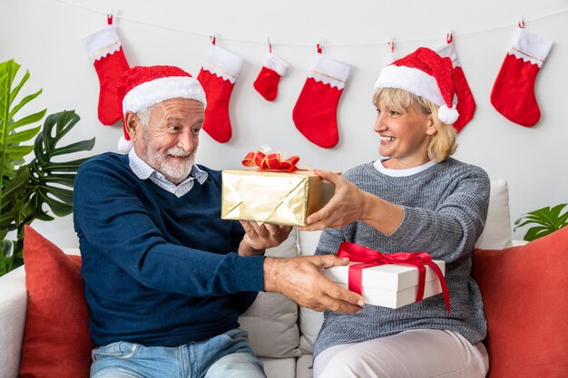 Senior couple husband and wife exchange giving gift present sitting on sofa in room with Christmas tree and decoration