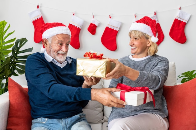 Senior couple husband and wife exchange giving gift present sitting on sofa in room with Christmas tree and decoration