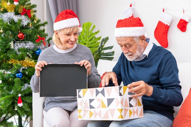 Senior couple husband and wife opening gift present box sitting on sofa in room with Christmas tree and decoration
