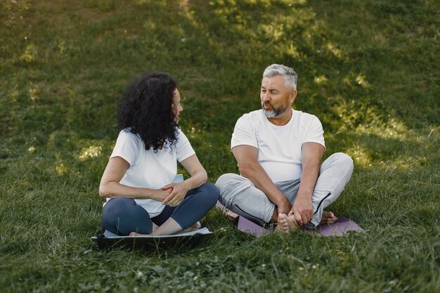 Senior couple is doing yoga outdoors. Stretching in park during sunrise. Brunette in a white t-shirt.