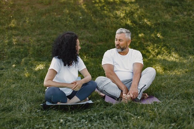 年配のカップルは屋外でヨガをやっています。日の出中に公園でストレッチします。白いtシャツのブルネット。