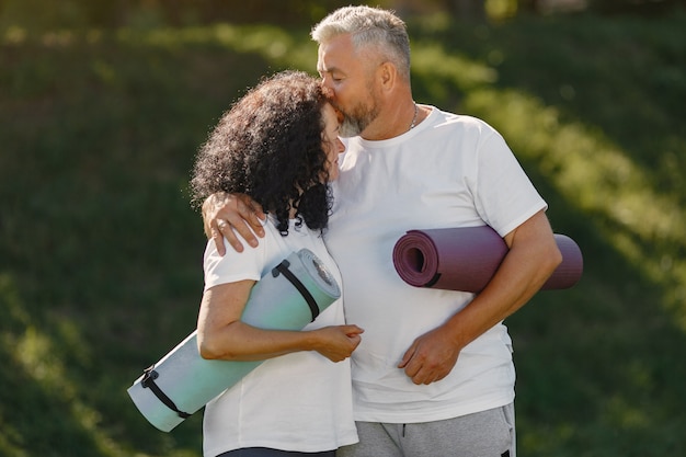 Senior couple is doing yoga outdoors. Stretching in park during sunrise. Brunette in a white t-shirt.