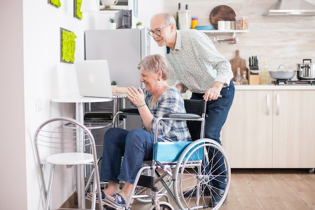 수석 부부는 부엌에서 태블릿 컴퓨터를 사용하여 손자와 화상 통화를 하는 동안 웃고 있습니다. 현대 통신 기술을 사용하는 마비된 장애인 노부인.
