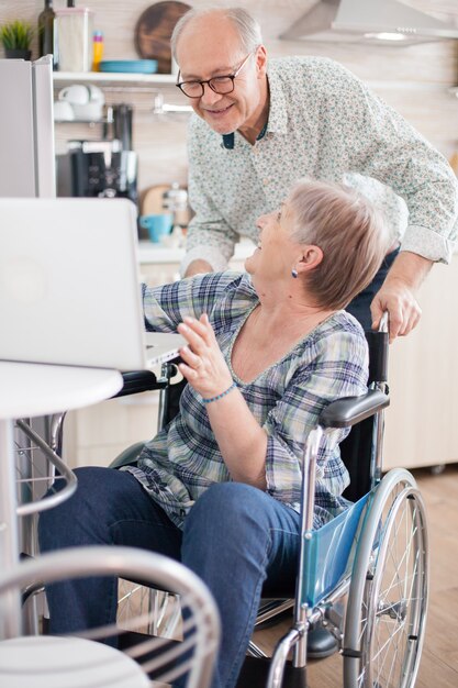 수석 부부는 부엌에서 태블릿 컴퓨터를 사용하여 손자와 화상 통화를 하는 동안 웃고 있습니다. 현대 통신 기술을 사용하는 마비된 장애인 노부인.
