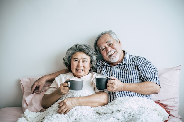 Пожилая пара смеется, выпивая кофе в спальне