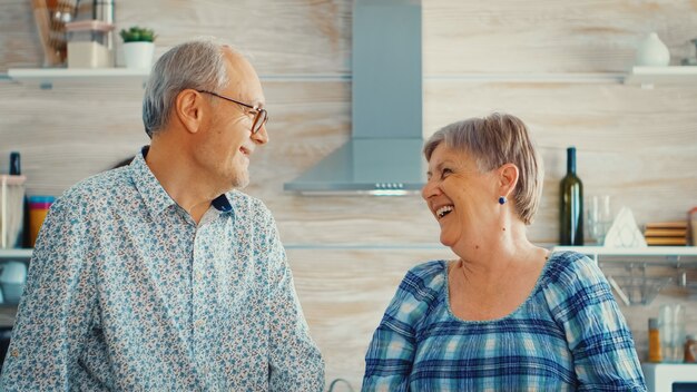 キッチンでカメラを見ながら笑っている年配のカップル。陽気な老人と女性の笑顔と笑い。生活を楽しんでいる居心地の良い家で幸せな高齢者退職者