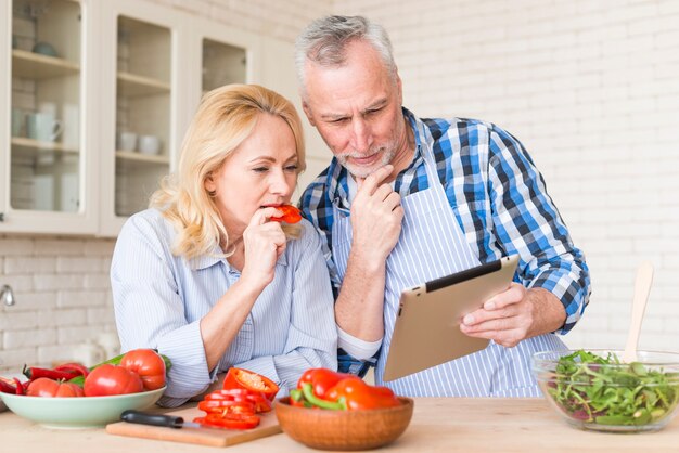 年配のカップルが台所で食べ物を準備しながらデジタルタブレットを見て