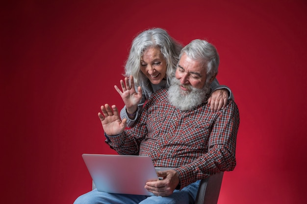 Foto gratuita coppie senior che esaminano computer portatile e che ondeggiano le loro mani contro il fondo rosso