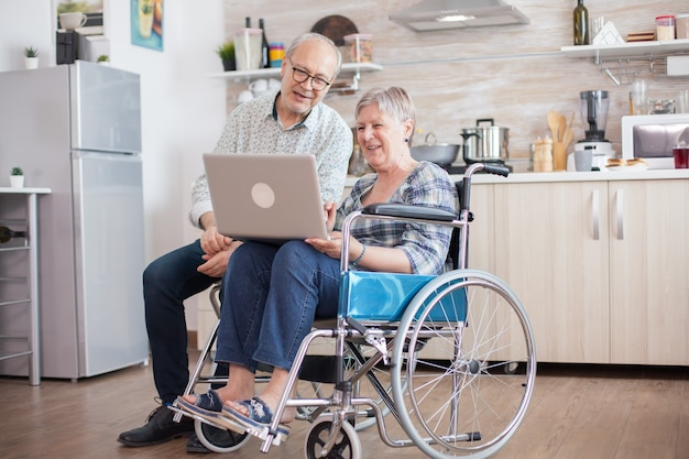 화상 통화 전에 웹캠을 보고 있는 수석 부부. 휠체어를 탄 장애인 노인과 남편이 부엌에서 태블릿 PC로 화상 회의를 하고 있습니다. 마비된 노부인과 그녀의 남편이