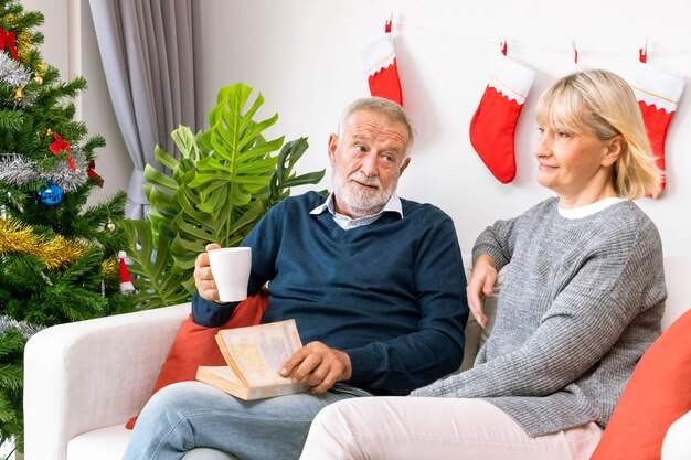 Senior couple man and woman reading a book and drinking coffee tea sitting on sofa with Christmas decoration in background