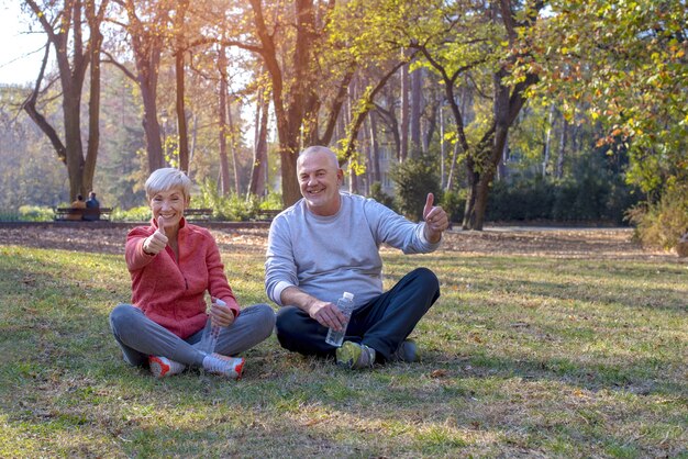 Senior couple sitting on the grass in a park happily, holding their thumbs up during daylight