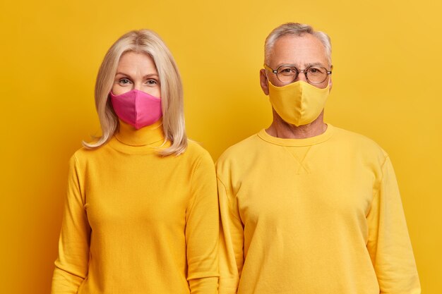 Senior couple stand closely to each other stay at home during quarantine wear protective face masks wear yellow clothing look seriously at front