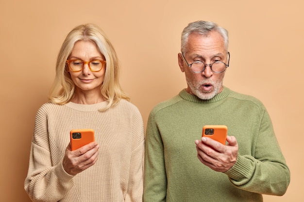 Пожилая пара стоит плечом к плечу, используя смартфоны для просмотра веб-сайта чтения сми в интернете, одетых в повседневные джемперы, изолированные на коричневой стене Бесплатные Фотографии