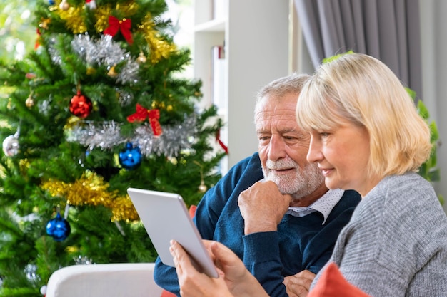 Пожилая пара использует планшет для видеозвонка, чтобы поприветствовать свою семью на рождественском фестивале, сидя на диване с украшением и елкой