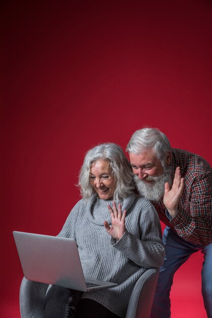 Video delle coppie senior che chiacchiera sul computer portatile che ondeggia le loro mani contro il fondo rosso