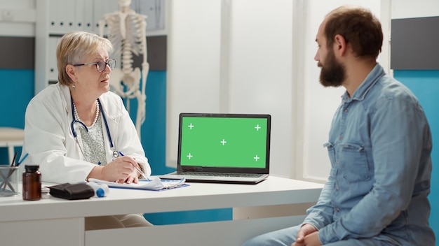 Senior doctor and patient looking at laptop with greenscreen at checkup visit. Man and woman using isolated template with blank chromakey background and mockup copyspace on display.