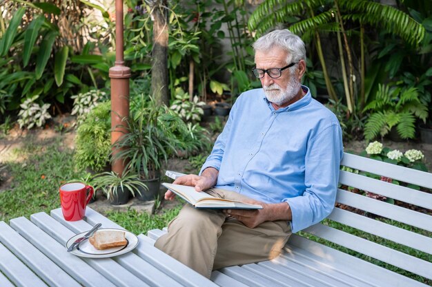 Senior elderly man reading book with breakfast and mug of coffee in garden