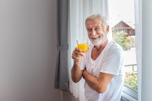 Senior elderly man standing at window in bedroom after waking up in morning with juice looking camera
