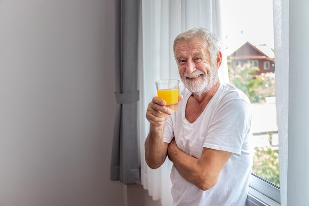 Senior elderly man standing at window in bedroom after waking up in morning with juice looking camera