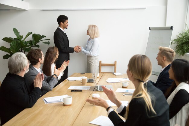 Senior female boss promoting handshaking african employee while team applauding