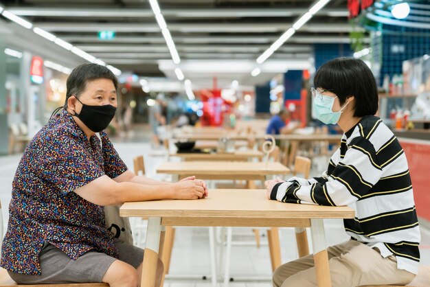 Senior female and young nephew sit talk conversation with distance new normal lifestyle after lockdown over from corona virus covid19 spread in mall department store