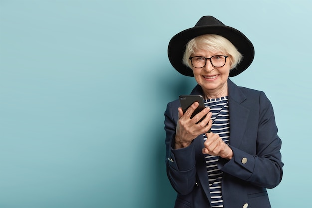 Senior glad woman has optical eyewear makes banking online on cell phone, uses modern technologies for searching information in internet, dressed in fashionable apparel, smiles friendly, isolated