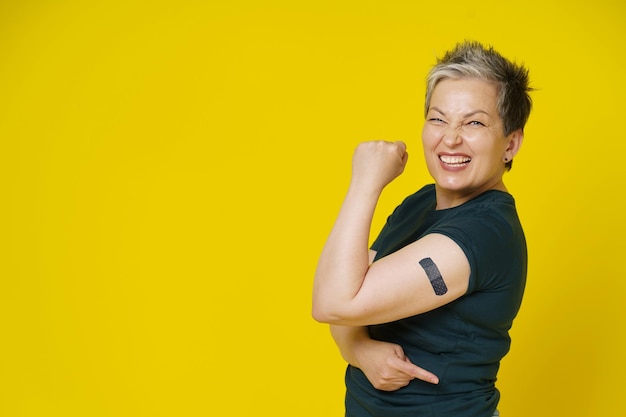 Senior grey haired woman rebelling showing nude shoulder with dark band aid or plaster on it isolated on yellow background Amazing mature woman 50s Vaccination and healthcare concept