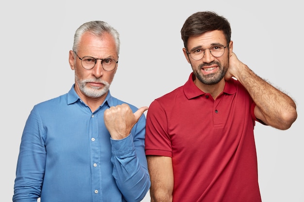 Senior grey haired young male with serious expression indicates with thumb at his young business partner who has nervous expression, stand closely to each other, isolated over white wall