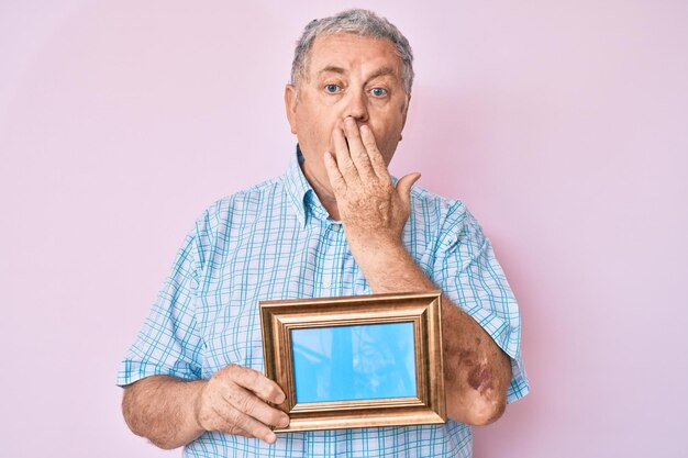 Free photo senior greyhaired man holding empty frame covering mouth with hand shocked and afraid for mistake surprised expression