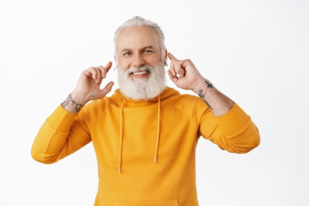 Senior hipster guy with tattoos listen music in wireless earphones, touching his headphones in ears and smiling pleased, enjoy awesome sound of earbuds, white wall