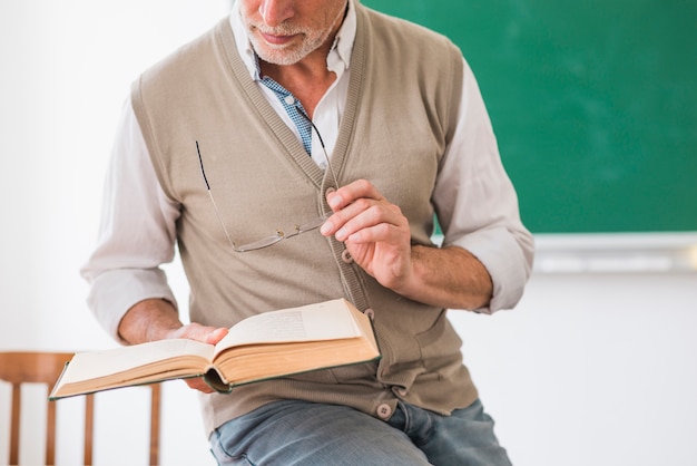 senior male professor holding book and glasses in classroom