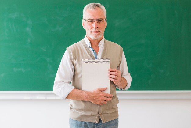 Senior male professor holding notebook standing against blackboard