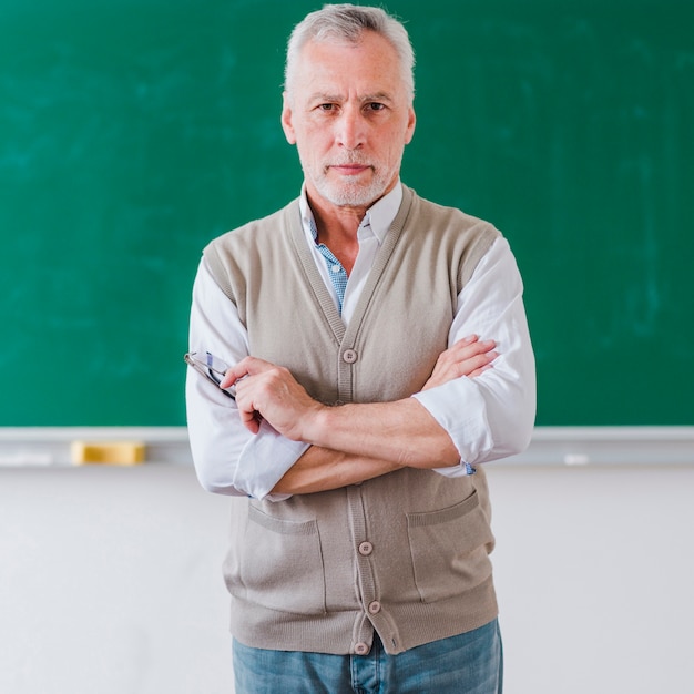 Senior male professor with arms crossed standing against chalkboard