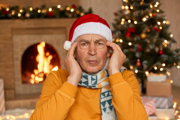 Senior male in Santa hat looking at camera and keeping hands on temples