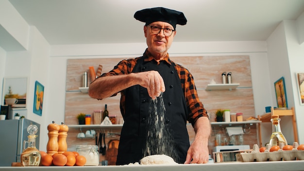 Free photo senior man adding flour on dough by hand looking at camera smiling. retired elderly chef with bonete and uniform sprinkling, sieving spreading rew ingredients with hand baking homemade pizza and bread