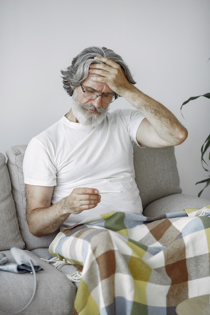 Senior man alone sitting on sofa. Sick man covered with plaid. Grangfather with thermometer.