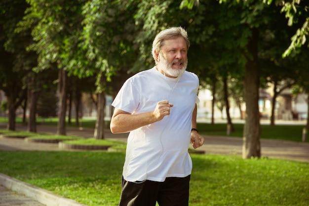 Senior man as runner with armband or fitness tracker at the city's street. Caucasian male model practicing jogging and cardio trainings in summer's morning. Healthy lifestyle, sport, activity concept.