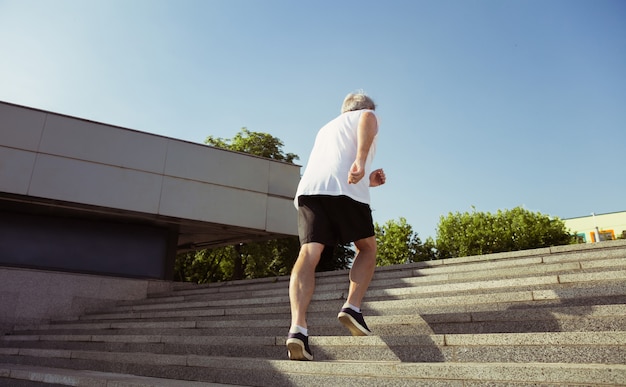 街の通りで腕章またはフィットネストラッカーを持つランナーとしての年配の男性。夏の朝にジョギングとカーディオトレーニングを練習している白人男性モデル。健康的なライフスタイル、スポーツ、活動の概念。