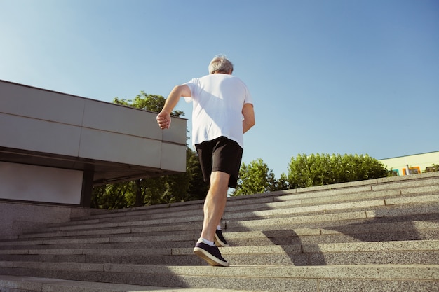 Senior man as runner with armband or fitness tracker at the city's street. Caucasian male model practicing jogging and cardio trainings in summer's morning. Healthy lifestyle, sport, activity concept.