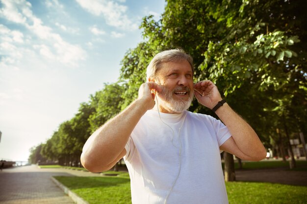 Senior man as runner with fitness tracker at the city's street. Caucasian male model using gadgets while jogging and cardio training in summer's morning. Healthy lifestyle, sport, activity concept.