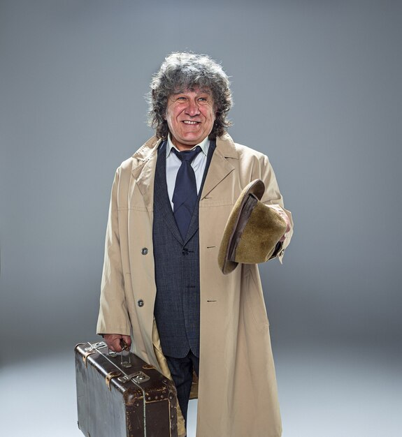 The senior man in cloak as detective or mafia boss. Studio shot on gray in retro stile. Mature man with hat and suitcase