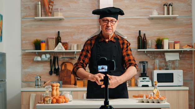 Senior man creating content for culinary blog, explaining the recipe step by step. Retired blogger chef influencer using internet technology communicating on social media with digital equipment