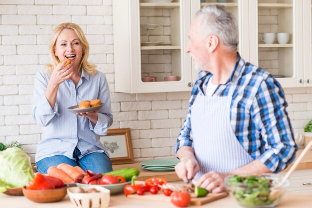 Старший мужчина нарезка овощей на разделочную доску, глядя на ее жена ест кексы на кухне