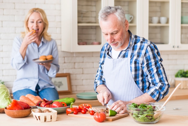 Старший мужчина резки овощей на разделочную доску с женой, ест кексы на фоне на кухне