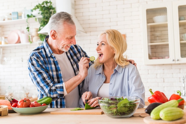 Пожилой мужчина кормит кусочек огурца своей жене на кухне