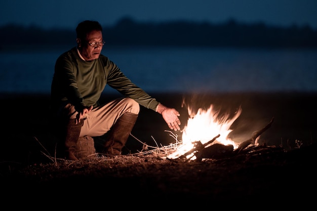 早朝の寒い時期に焚き火の炎で手を温める湖の近くに座っている眼鏡をかけた年配の男性、コピースペース