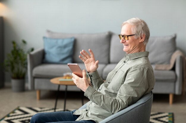 Старший мужчина разговаривает по видеосвязи с помощью планшета, сидящего на стуле в гостиной