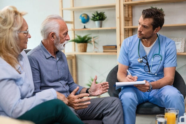 Senior man and his wife explaining their health issues to a male doctor visiting them at home and taking notes